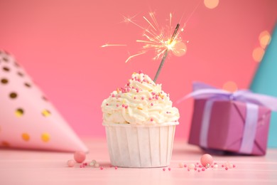 Photo of Burning sparkler in cupcake, sprinkles and party decor on pink table against color background with blurred lights