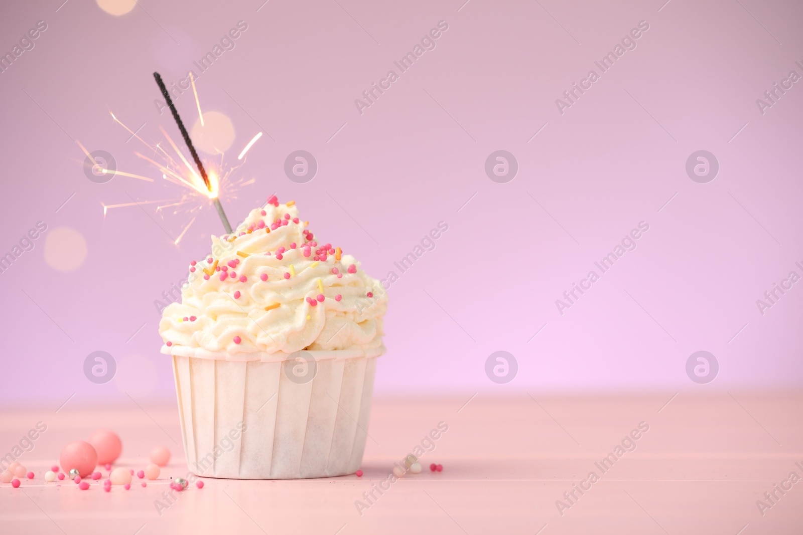 Burning sparkler in cupcake and sprinkles on pink table against violet background with blurred lights, space for text Photo of Burning sparkler in cupcake and sprinkles on pink table against violet background with blurred lights, space for text