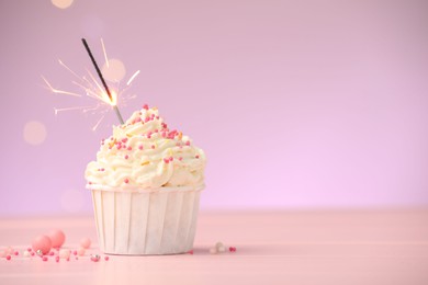 Photo of Burning sparkler in cupcake and sprinkles on pink table against violet background with blurred lights, space for text