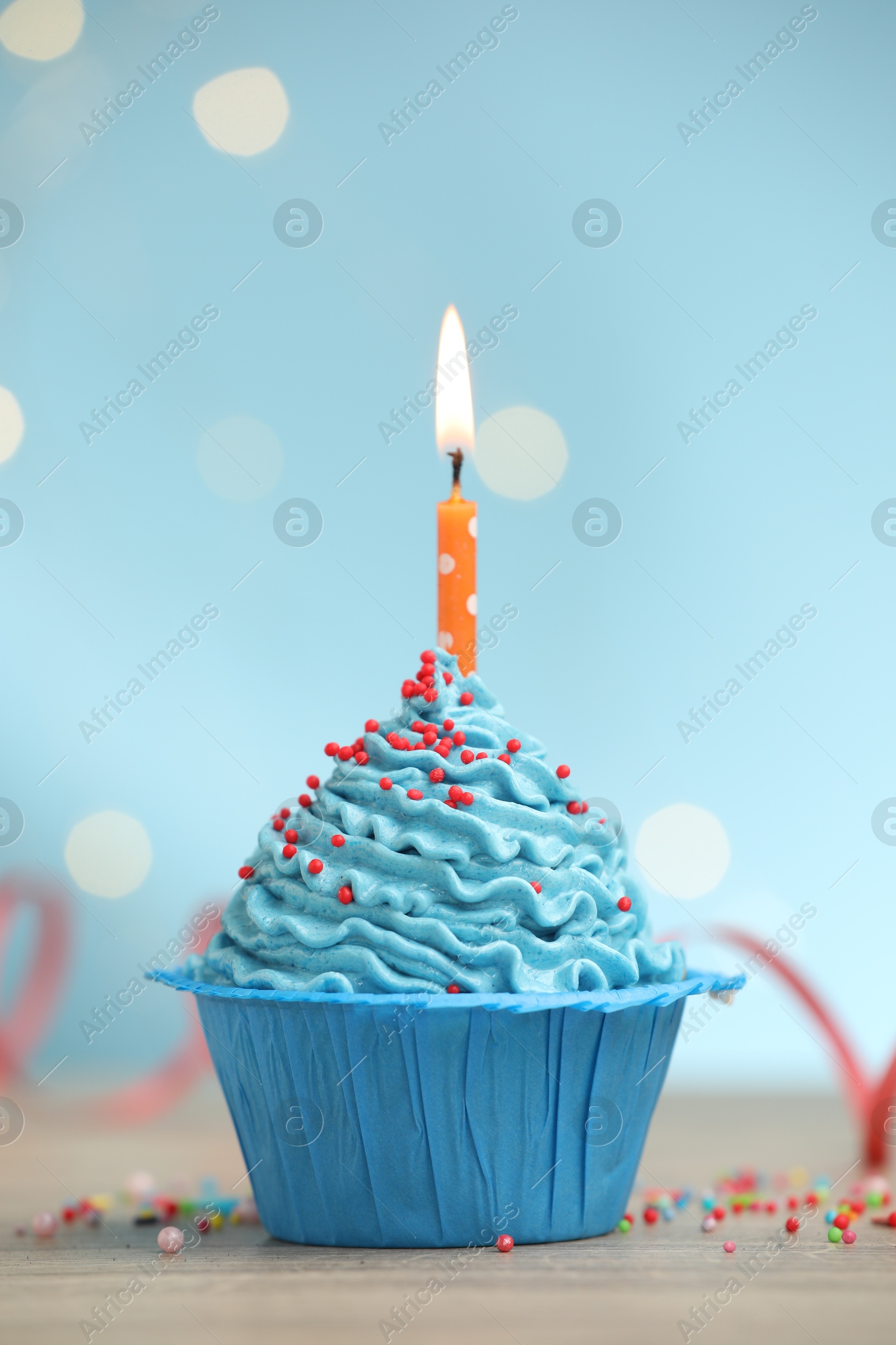 Burning candle in cupcake and sprinkles on wooden table against light blue background with blurred lights Photo of Burning candle in cupcake and sprinkles on wooden table against light blue background with blurred lights