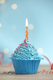 Photo of Burning candle in cupcake and sprinkles on wooden table against light blue background with blurred lights