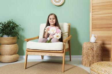 Photo of Cute little girl with bouquet of beautiful peonies in armchair near green wall indoors