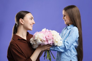 Photo of Cute little girl giving her mother bouquet of beautiful peonies on purple background