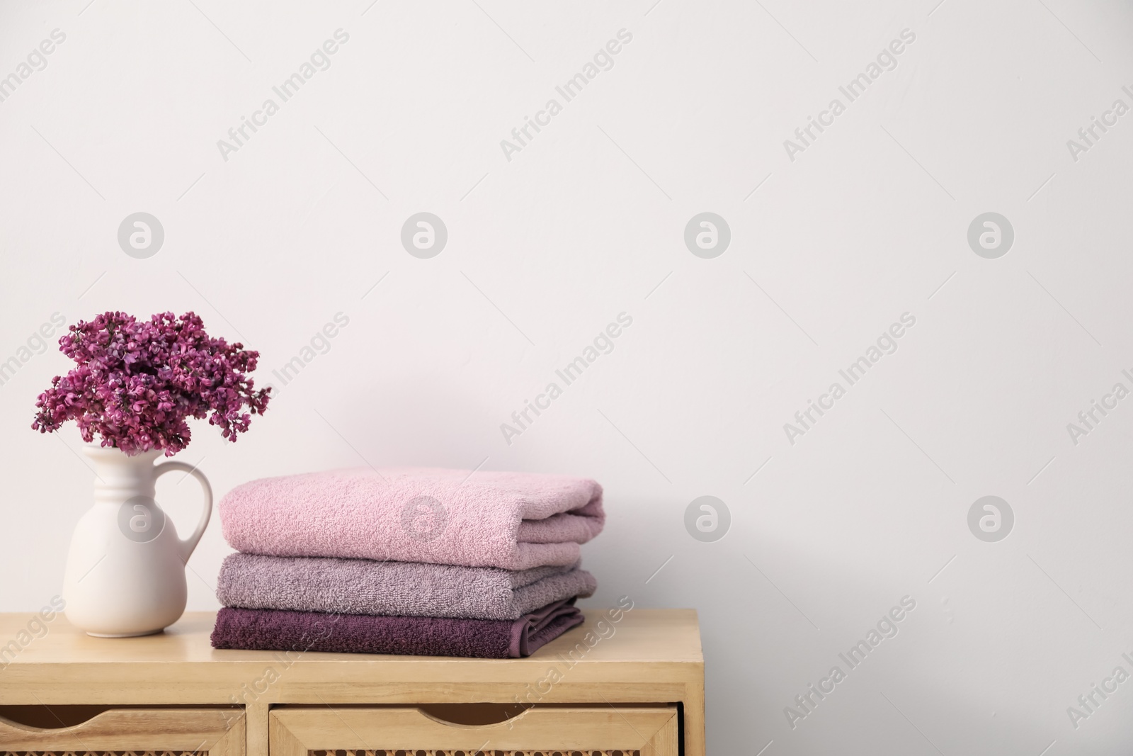 Stack of soft towels and lilac flowers on wooden cabinet near white wall indoors, space for text Photo of Stack of soft towels and lilac flowers on wooden cabinet near white wall indoors, space for text