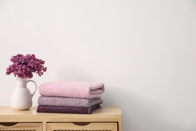 Photo of Stack of soft towels and lilac flowers on wooden cabinet near white wall indoors, space for text