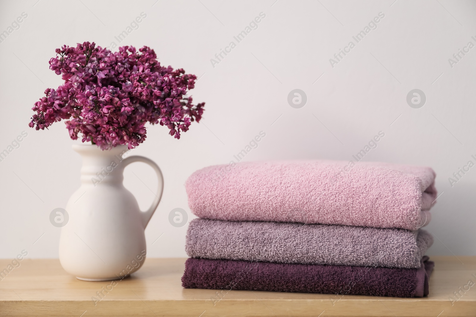 Stack of soft towels and lilac flowers on wooden table near white wall indoors Photo of Stack of soft towels and lilac flowers on wooden table near white wall indoors