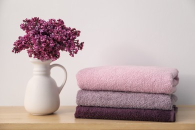 Photo of Stack of soft towels and lilac flowers on wooden table near white wall indoors