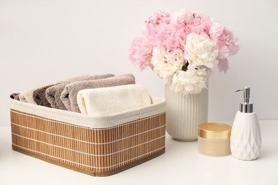 Photo of Soft towels in storage basket, peony flowers and cosmetic products on white table indoors