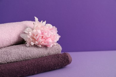 Photo of Stack of soft towels and peony flower on purple background, closeup. Space for text