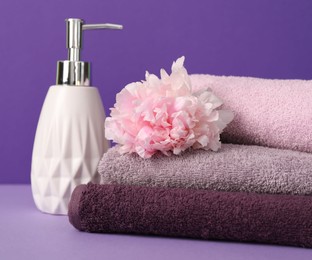 Photo of Stack of soft towels, peony flower and liquid soap on purple background, closeup