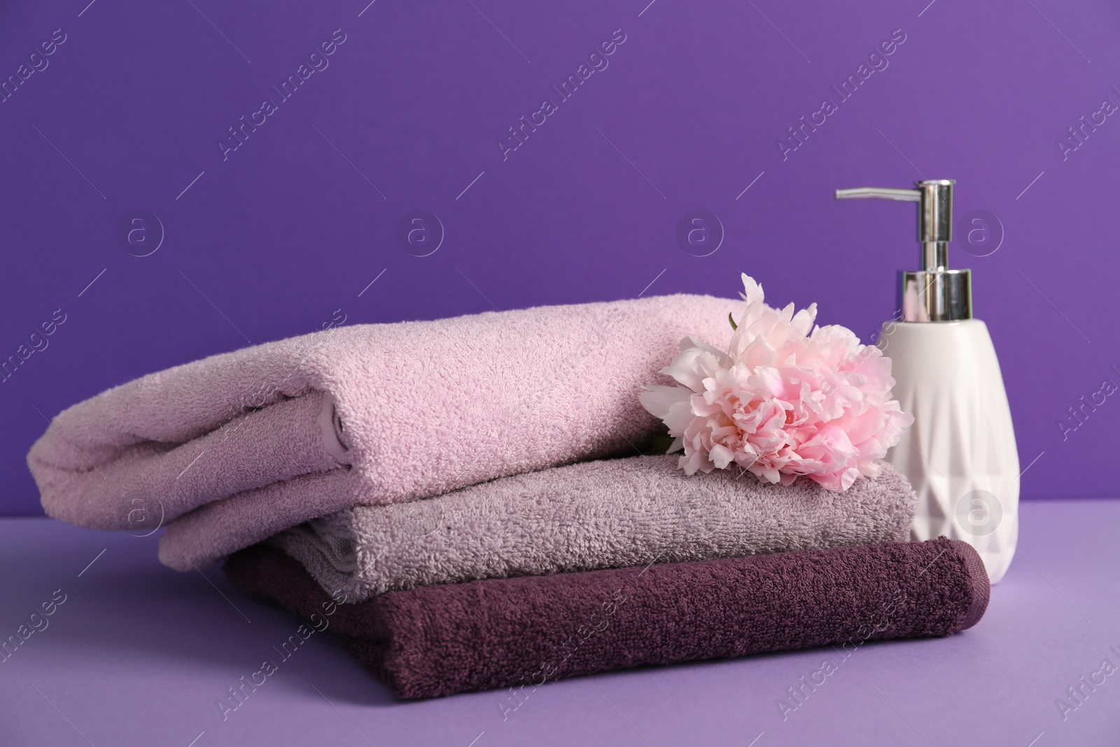 Stack of soft towels, peony flower and liquid soap on purple background Photo of Stack of soft towels, peony flower and liquid soap on purple background