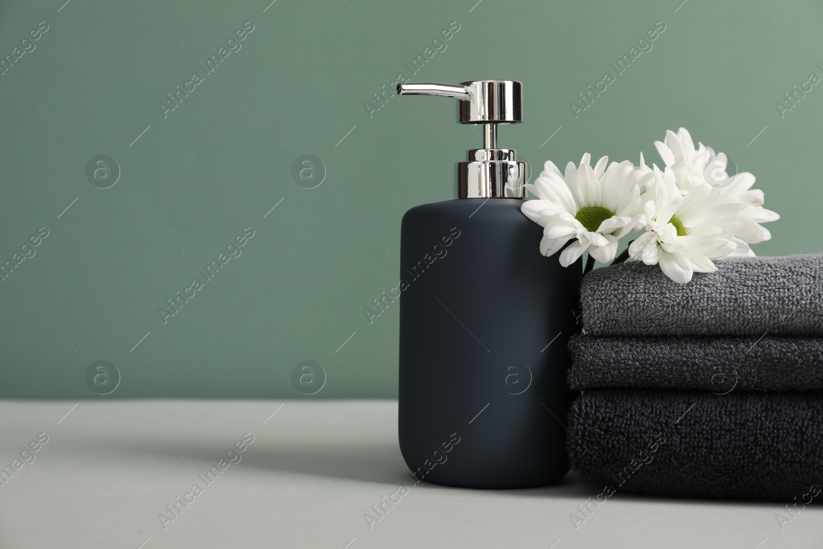Stack of soft towels, liquid soap and chrysanthemum flowers on table against greyish green background, closeup. Space for text Photo of Stack of soft towels, liquid soap and chrysanthemum flowers on table against greyish green background, closeup. Space for text