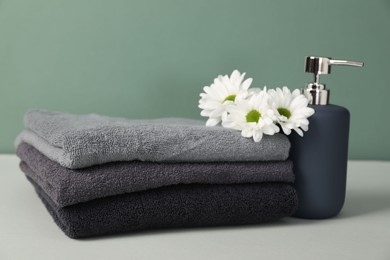 Photo of Stack of soft towels, liquid soap and chrysanthemum flowers on table against greyish green background, closeup