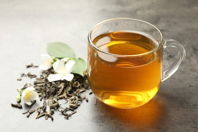 Photo of Aromatic jasmine tea in glass cup, brew and flowers on grey table, closeup