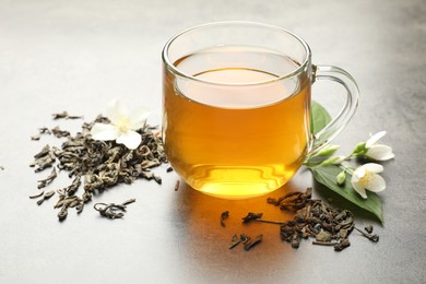 Photo of Aromatic jasmine tea in glass cup, brew and flowers on grey table, closeup