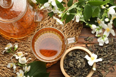 Photo of Aromatic jasmine tea in glass cup, teapot, brew and flowers on wooden table, flat lay