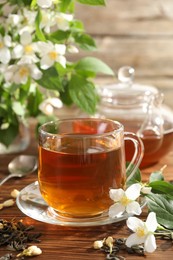 Photo of Aromatic jasmine tea in glass cup, teapot, brew and flowers on wooden table, closeup