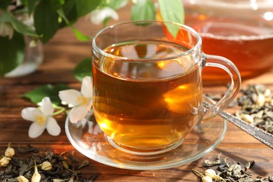 Photo of Aromatic jasmine tea in glass cup, brew and flowers on wooden table, closeup