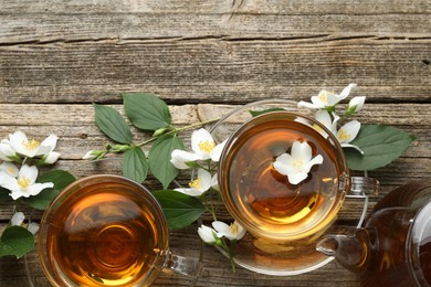Photo of Aromatic jasmine tea in glass cups, teapot, flowers and green leaves on wooden table, flat lay