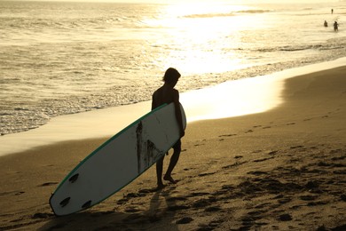 Photo of Bali, Indonesia - May 20, 2025: Man with surfboard walking along seashore at sunrise