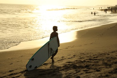 Photo of Bali, Indonesia - May 20, 2025: Man with surfboard walking along seashore at sunrise