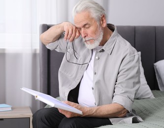 Photo of Senior man with vision problem reading book on bed at home
