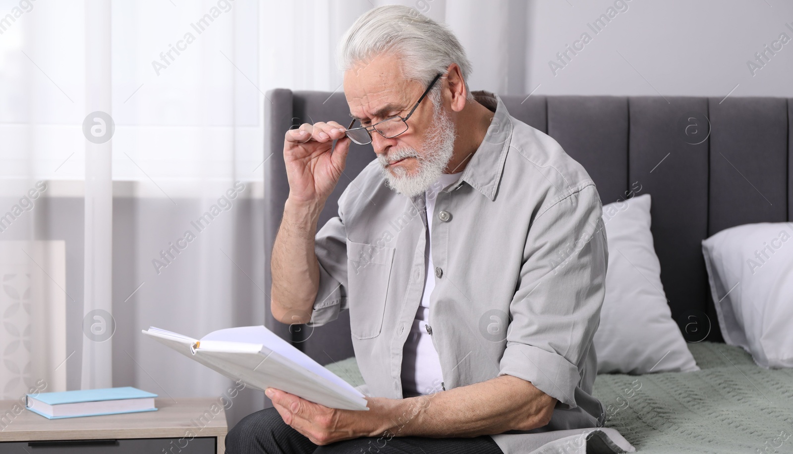 Photo of Senior man with vision problem reading book on bed at home