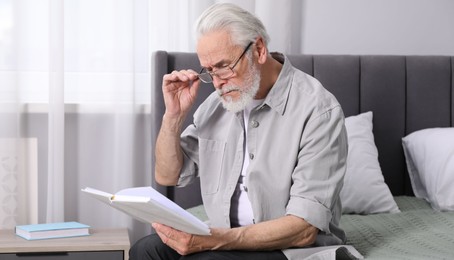 Photo of Senior man with vision problem reading book on bed at home