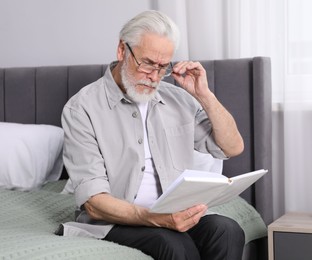 Photo of Senior man with vision problem reading book on bed at home