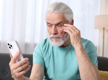 Photo of Senior man with vision problem using smartphone on sofa at home
