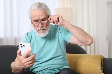 Photo of Senior man with vision problem using smartphone on sofa at home