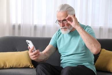 Photo of Senior man with vision problem using smartphone on sofa at home
