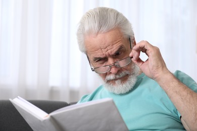 Photo of Senior man with vision problem reading book on sofa at home