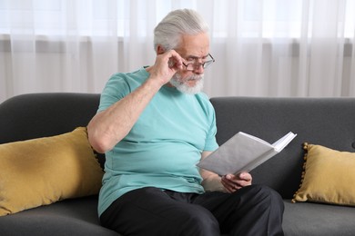 Photo of Senior man with vision problem reading book on sofa at home