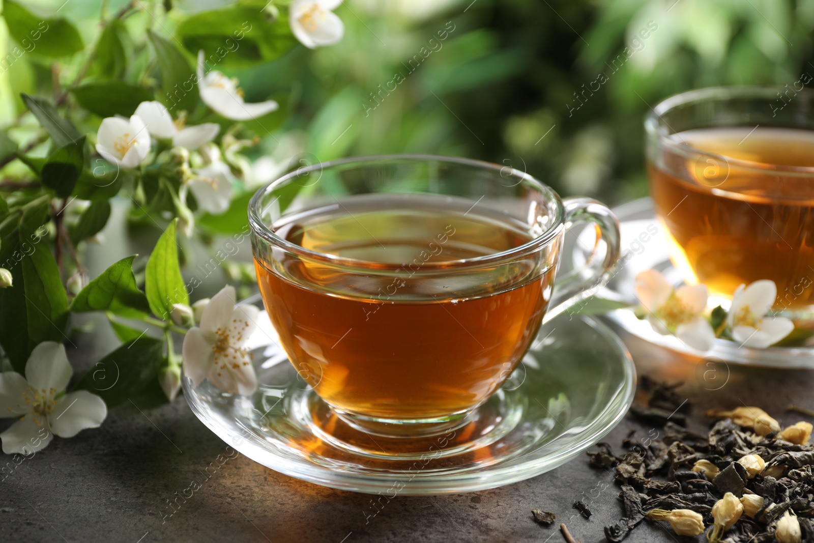 Photo of Aromatic jasmine tea in glass cups, brew and flowers on grey table outdoors, closeup