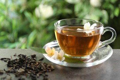 Photo of Aromatic jasmine tea in glass cup, brew and flowers on grey table outdoors, closeup
