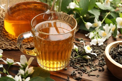 Photo of Aromatic jasmine tea in glass cup, teapot, brew and flowers on wooden table, closeup