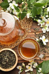 Photo of Aromatic jasmine tea in glass cup, teapot, brew and flowers on wooden table, flat lay