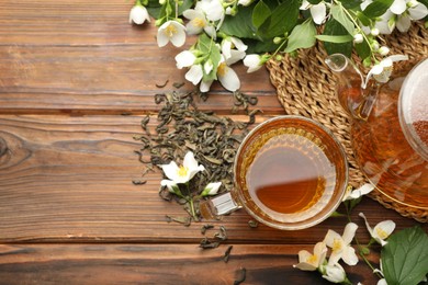 Photo of Aromatic jasmine tea in glass cup, teapot, brew and flowers on wooden table, flat lay. Space for text