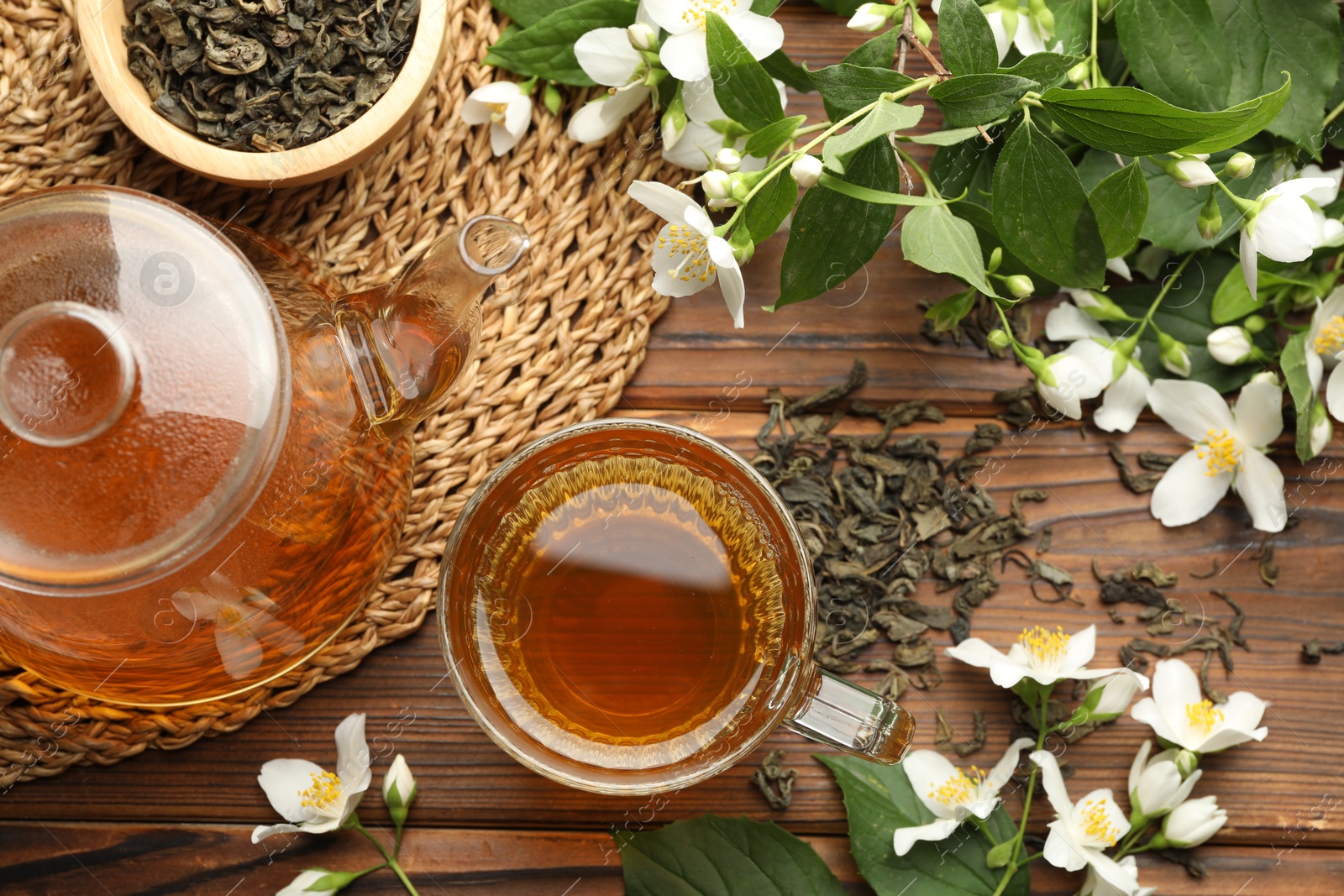 Photo of Aromatic jasmine tea in glass cup, teapot, brew and flowers on wooden table, flat lay