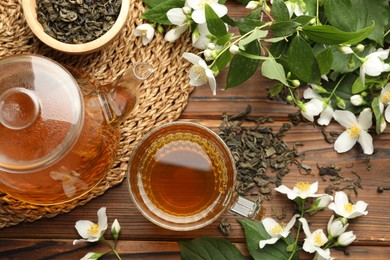 Photo of Aromatic jasmine tea in glass cup, teapot, brew and flowers on wooden table, flat lay