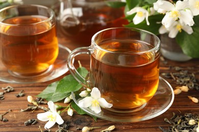 Photo of Aromatic jasmine tea in glass cups, teapot, brew and flowers on wooden table, closeup