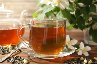 Photo of Aromatic jasmine tea in glass cup, teapot, brew and flowers on wooden table, closeup