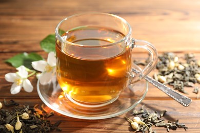 Photo of Aromatic jasmine tea in glass cup, brew and flowers on wooden table, closeup