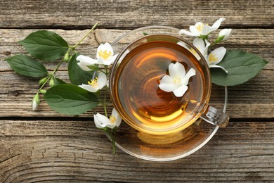 Photo of Aromatic jasmine tea in glass cup, flowers and green leaves on wooden table, flat lay