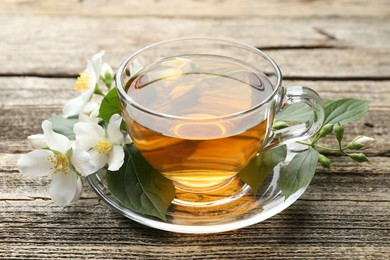 Photo of Aromatic jasmine tea in glass cup, flowers and green leaves on wooden table, closeup