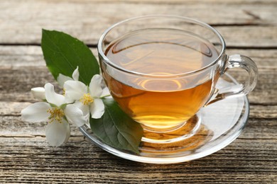 Photo of Aromatic jasmine tea in glass cup, flowers and green leaves on wooden table, closeup