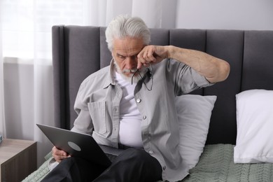 Photo of Senior man with vision problem using laptop on bed at home