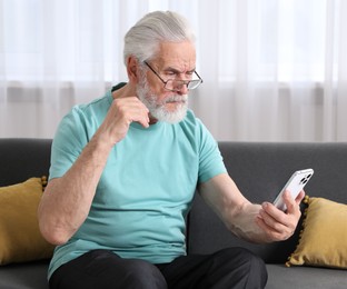 Photo of Senior man with vision problem using smartphone on sofa at home