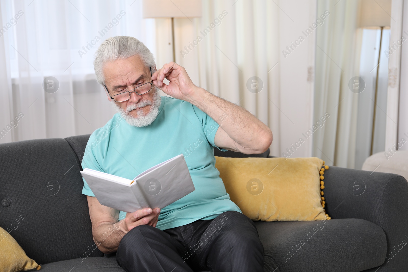 Photo of Senior man with vision problem reading book on sofa at home